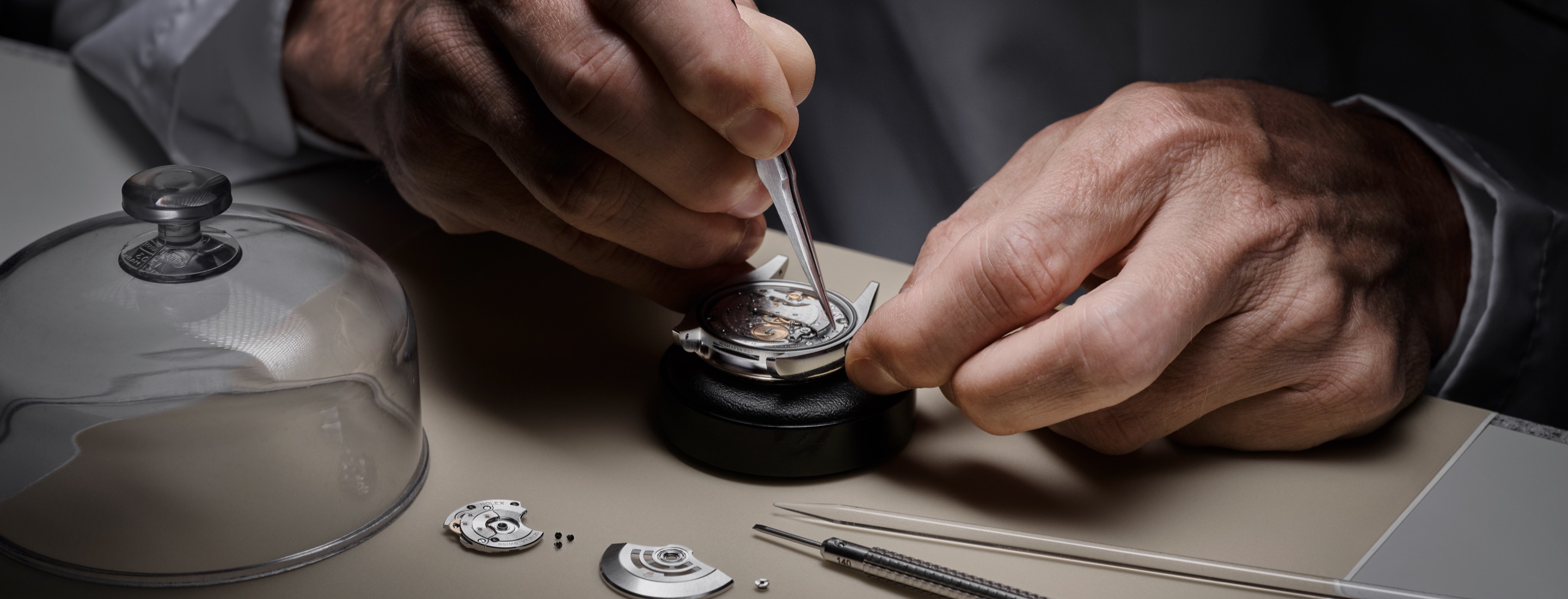 Rolex Uhrmacher zerlegt ein Uhrwerk an der Werkbank unter der Glasglocke
